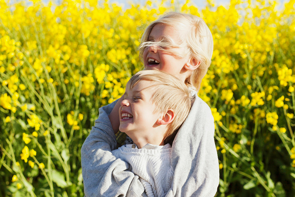 Foto på två barn som håller om varandra framför ett gult rapsfält. 