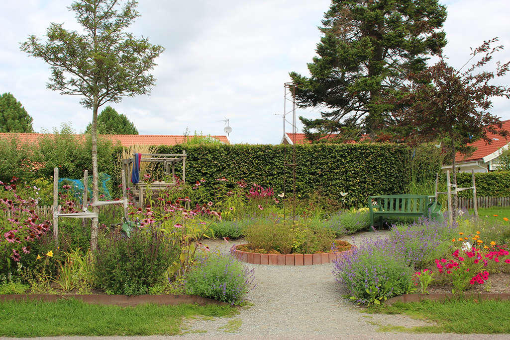 Foto på en av idéträdgårdarna längs med Åpromenaden. I bilden syns en liten fickpark med färgglada blommor, parkmöbler och grusgång.